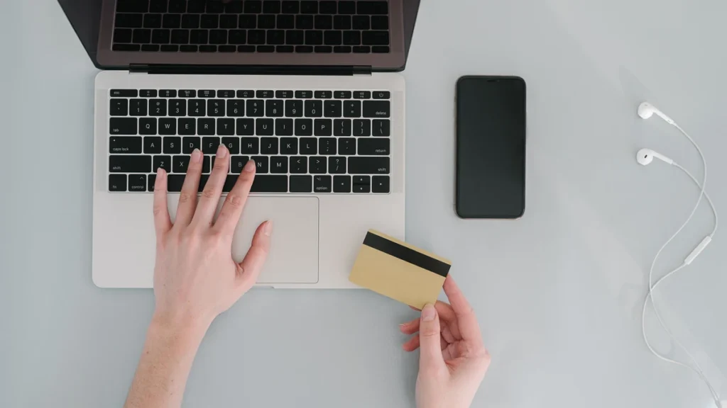Hands using a laptop while holding a bank card beside a smartphone.