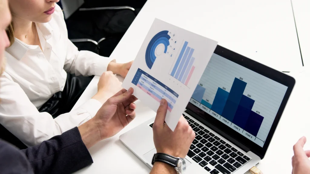 Business professionals reviewing printed charts beside a laptop displaying a bar graph.