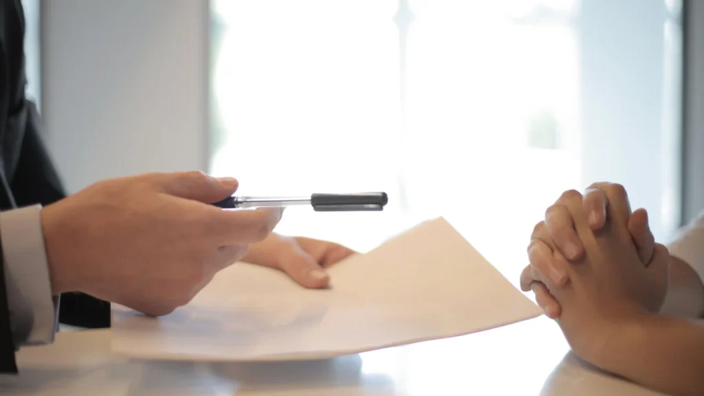 A person handing a pen and contract documents to another person across a desk.