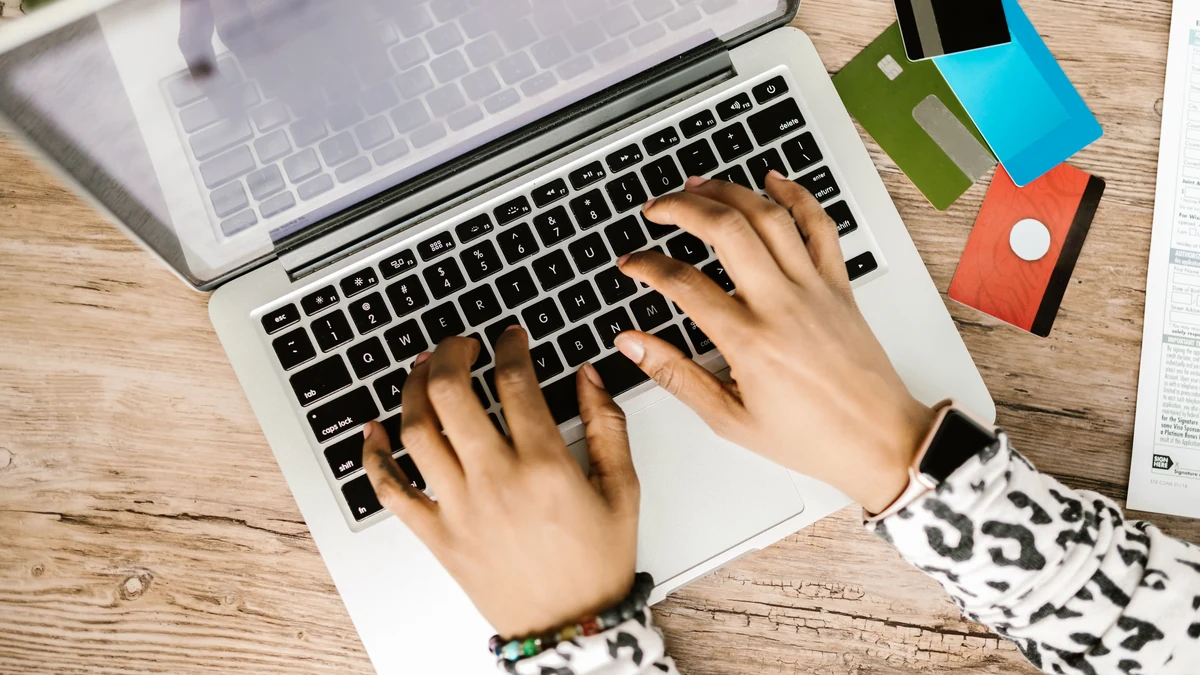 Hands using a laptop beside several credit cards on a wooden desk.