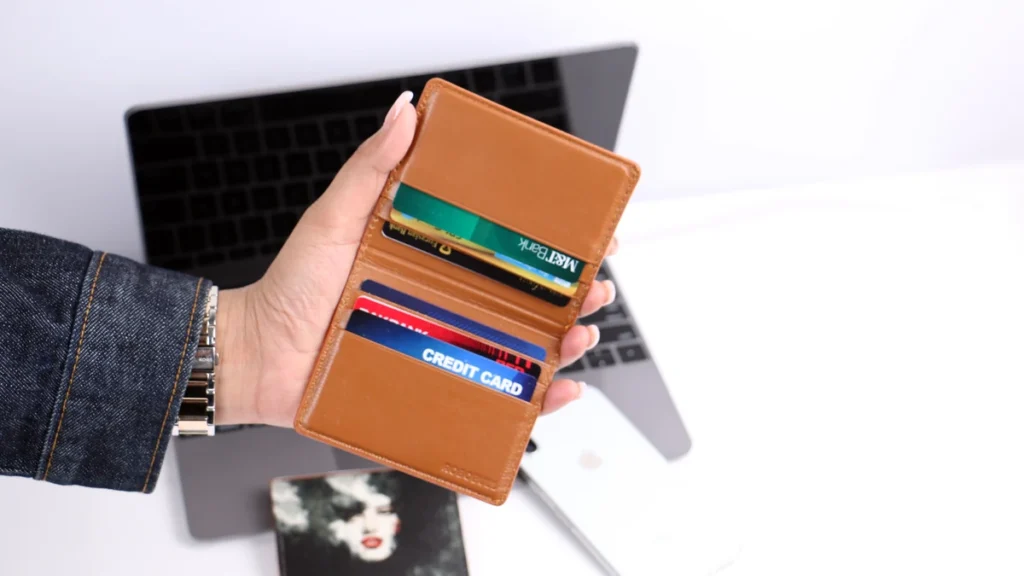 Hand holding a brown wallet containing several credit cards in front of a laptop.