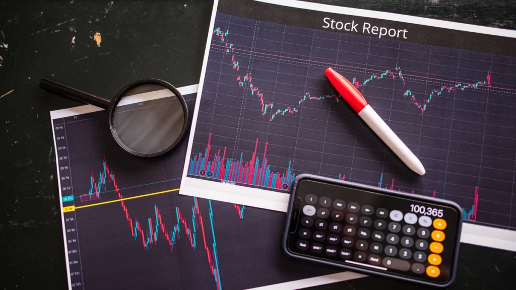 Printed stock charts, a magnifying glass and a calculator on a dark table.