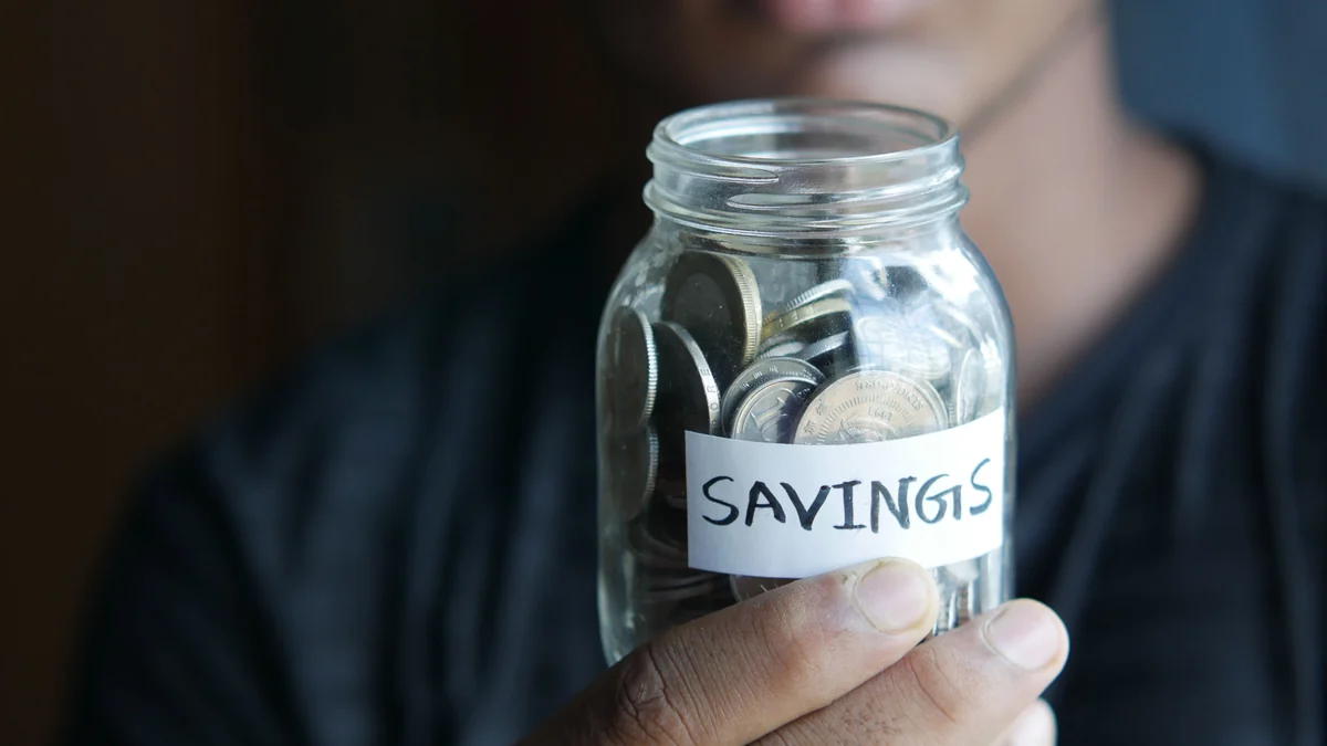 A person holding a glass jar of coins labeled Savings.