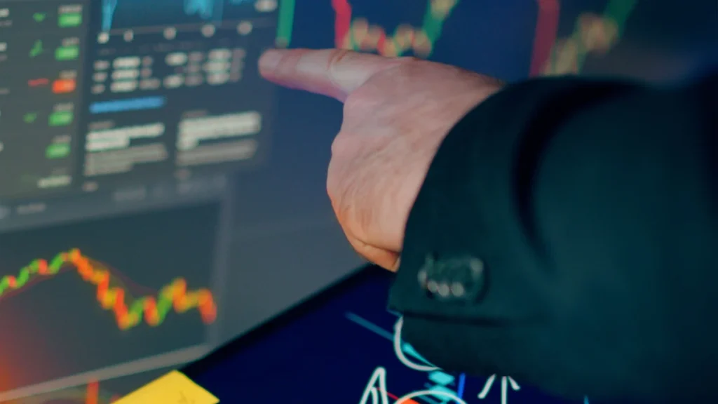 A person pointing at stock market charts displayed on tablet and computer screens.