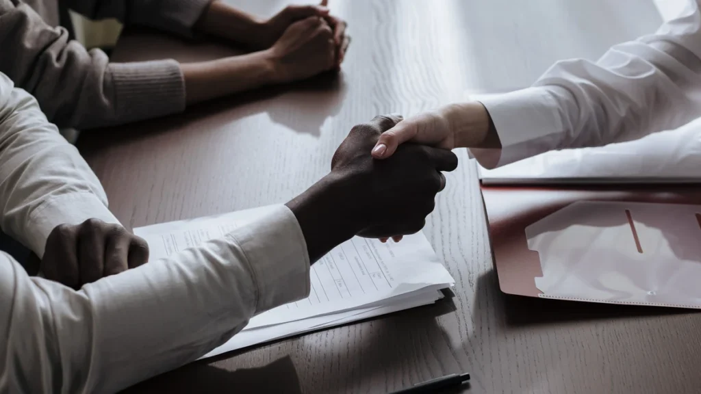Two people shaking hands over paperwork at a desk.