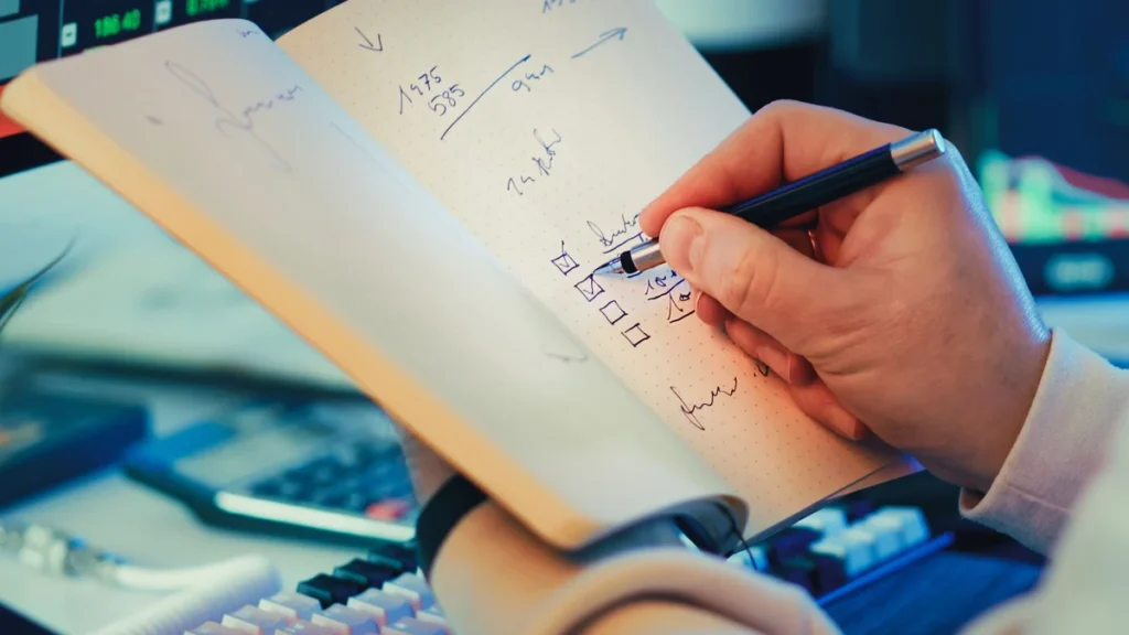 A person writing notes while reviewing stock market information on computer screens.
