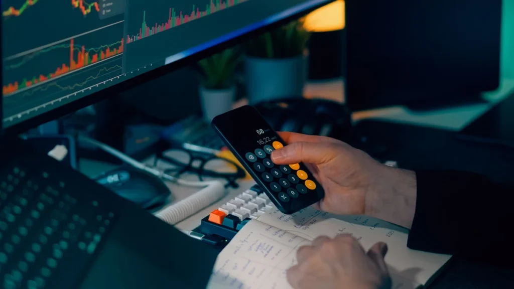 A person using a calculator while reviewing financial market charts at a trading workstation.