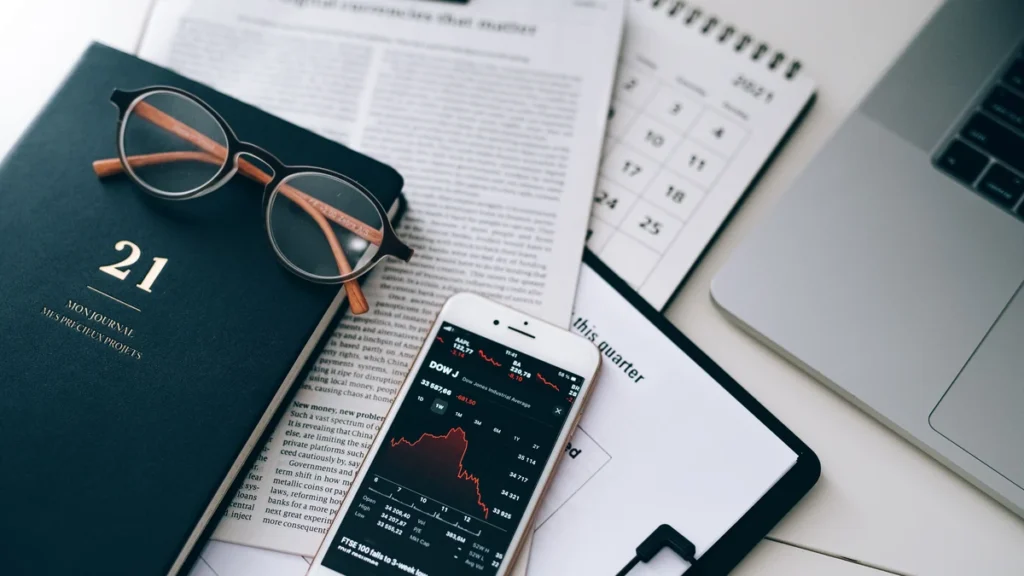 Smartphone displaying a falling market chart beside financial documents, a notebook and a laptop.