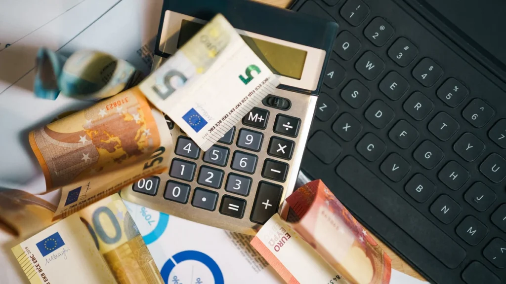 Calculator, euro banknotes, financial documents and a laptop keyboard on a desk.