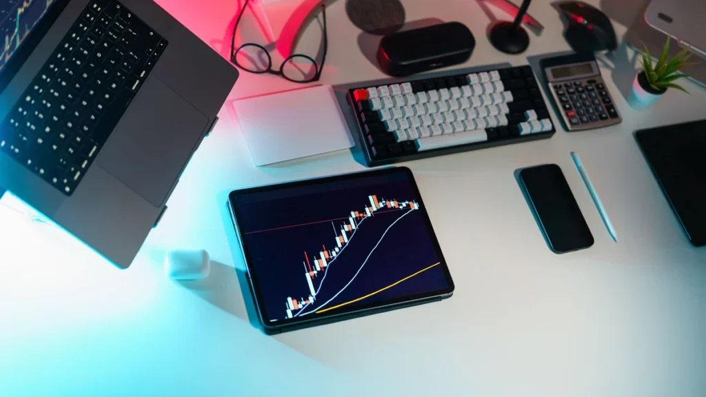 A financial trading workspace with a tablet displaying a market chart beside a laptop, keyboard and calculator.
