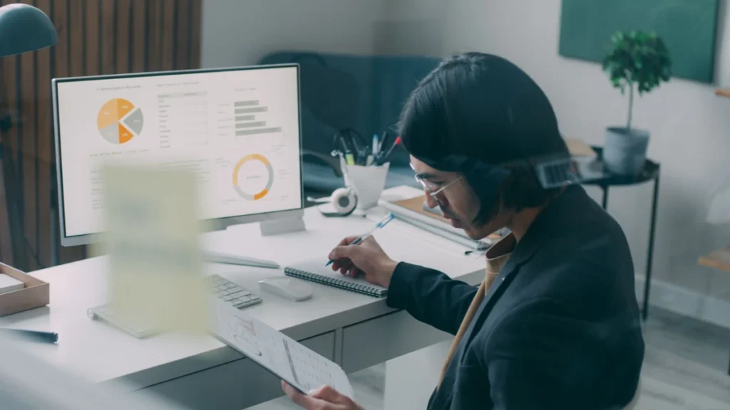 A business professional reviewing charts on a computer and printed report at an office desk.