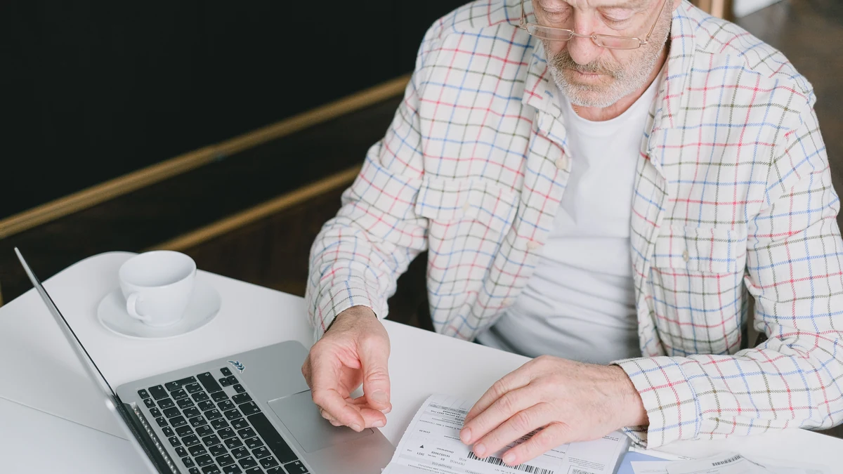 Older man reviewing financial documents beside a laptop at a table.