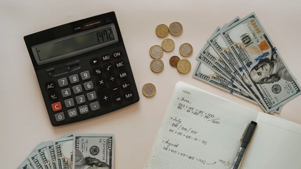 Calculator, cash, coins and a notebook with handwritten budget calculations on a desk.
