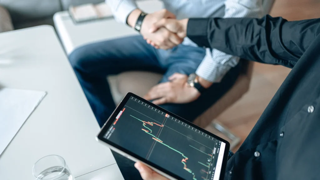 Two business professionals shaking hands while reviewing a financial market chart on a tablet.