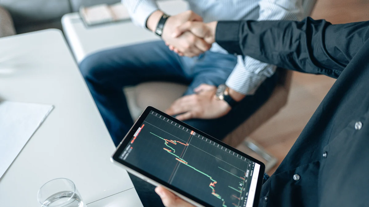 Two business professionals shaking hands while reviewing a financial market chart on a tablet.