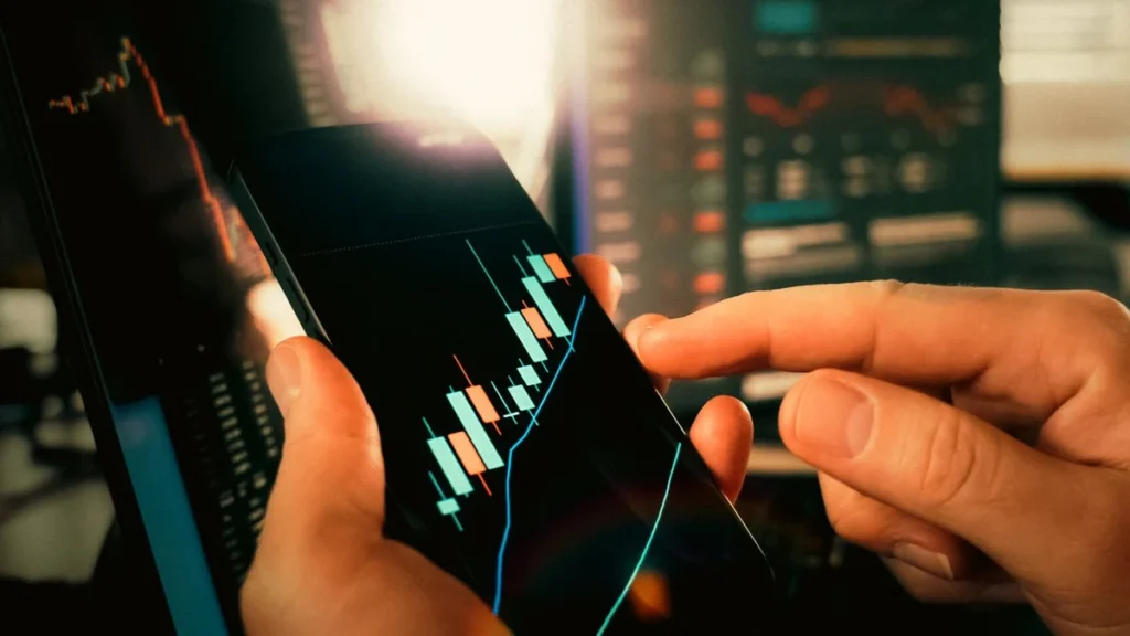 Hands holding a smartphone showing financial market candlestick charts in front of trading screens.
