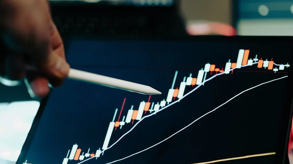 Digital tablet displaying a financial market candlestick chart with blurred trading screens in the background.
