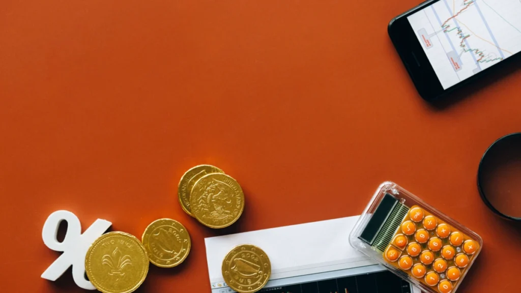 Gold-colored coins placed beside a calculator and market charts on a red background.