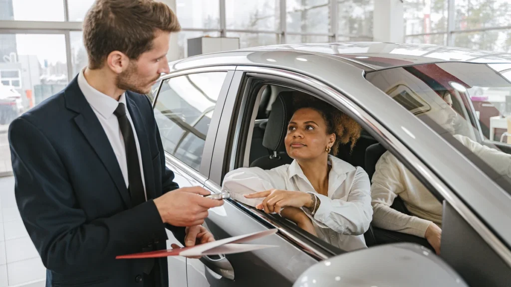 A customer sitting in a car receives the keys from a dealership representative.