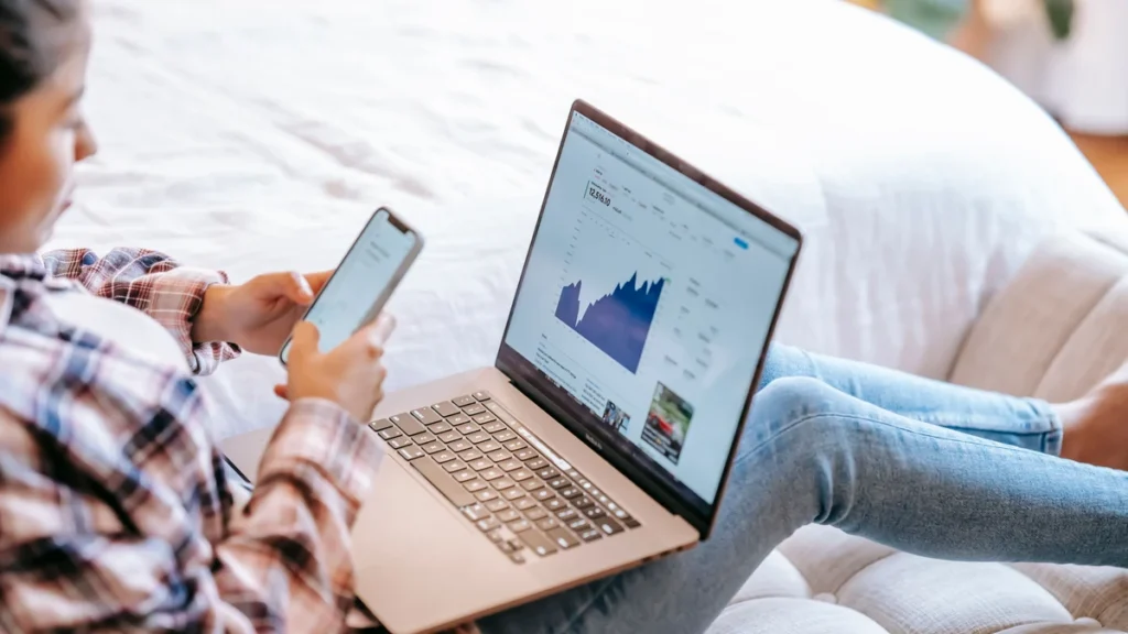 An investor reviewing market data on a laptop while holding a smartphone.