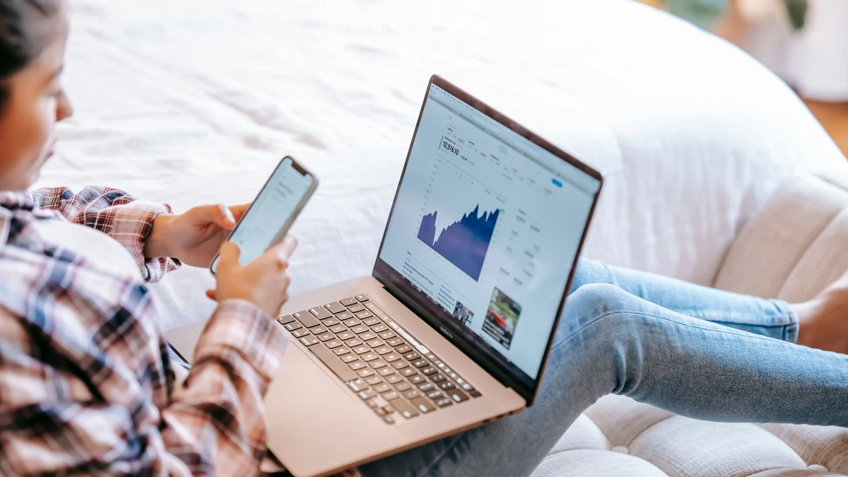 An investor reviewing market data on a laptop while holding a smartphone.