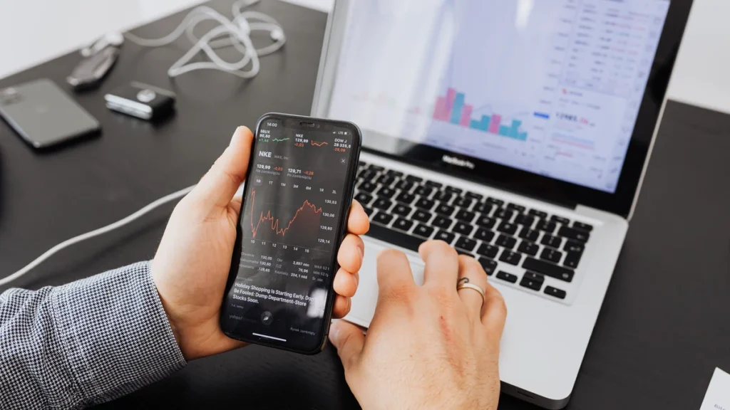 A person viewing stock market charts on a smartphone beside a laptop displaying financial market data.