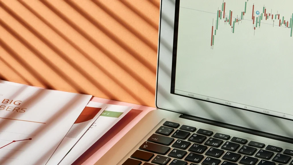 A hand using a laptop displaying a financial market chart beside printed reports.