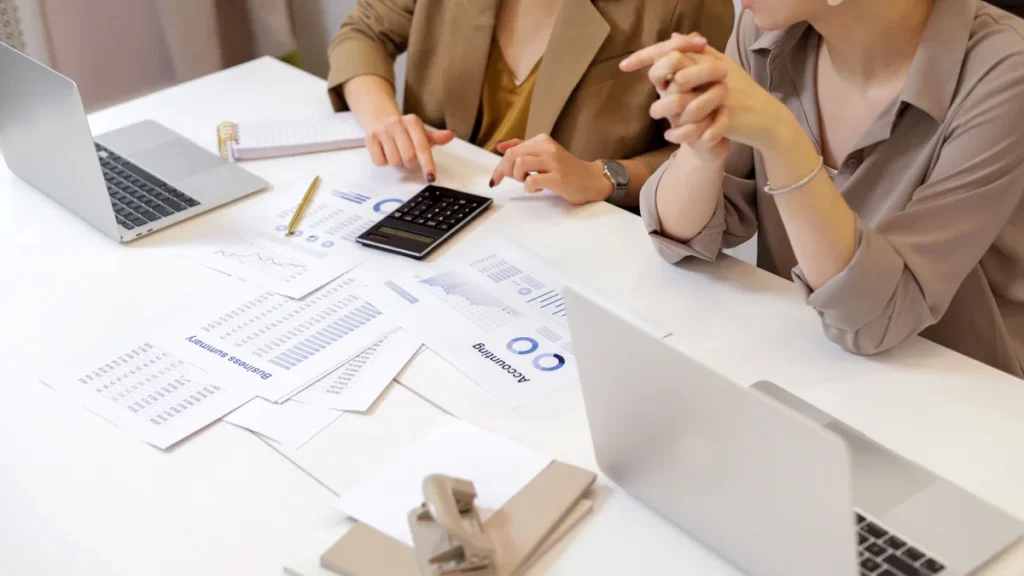 Two people reviewing financial charts and figures with a calculator and laptops at a desk.
