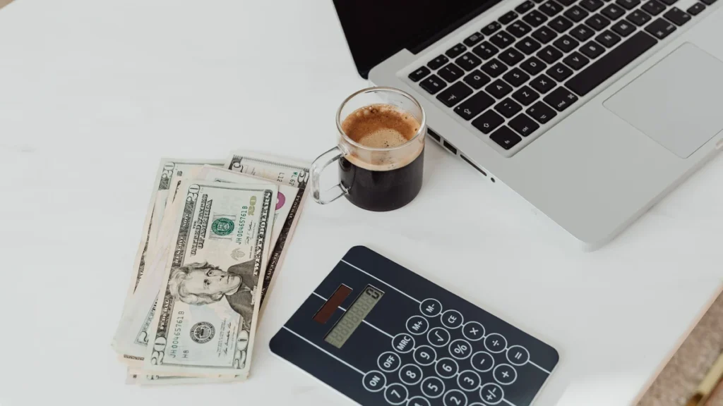 Calculator, laptop, coffee and U.S. dollar bills arranged on a white desk.