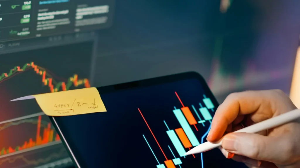 Hands using a stylus to analyze candlestick charts on a tablet in front of market screens.
