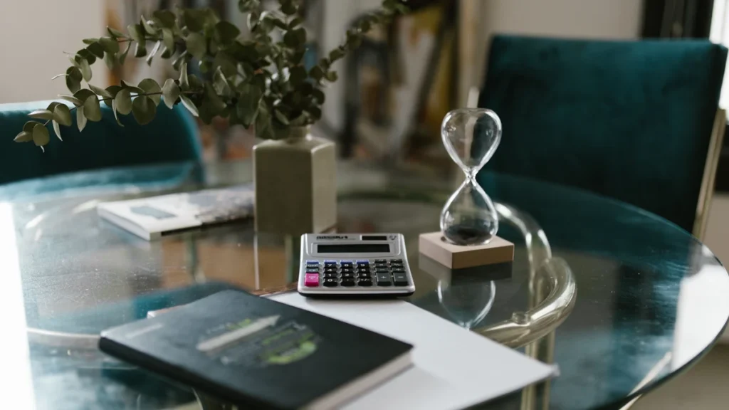 Hourglass and calculator on a glass table beside notebooks.