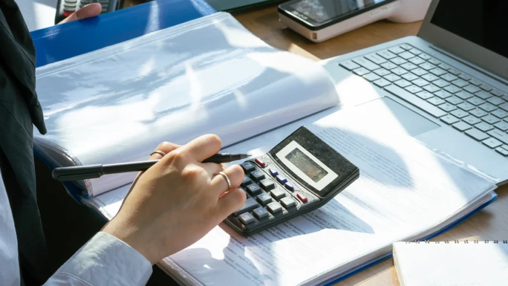 A person using a calculator while reviewing financial documents beside a laptop.