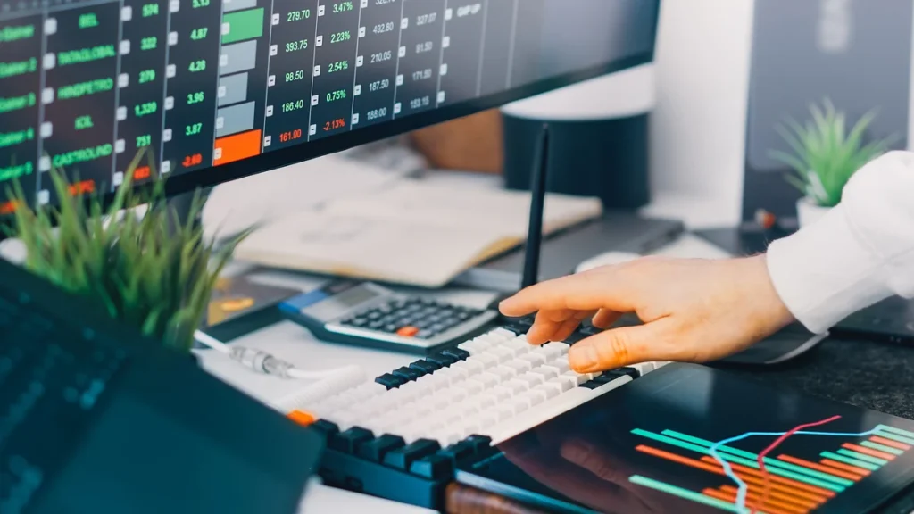 A person using a computer at a desk with market data and financial charts on screens.