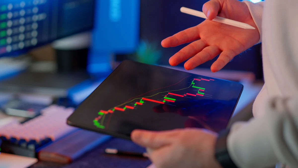 A person reviewing a financial candlestick chart on a tablet at a trading workstation.