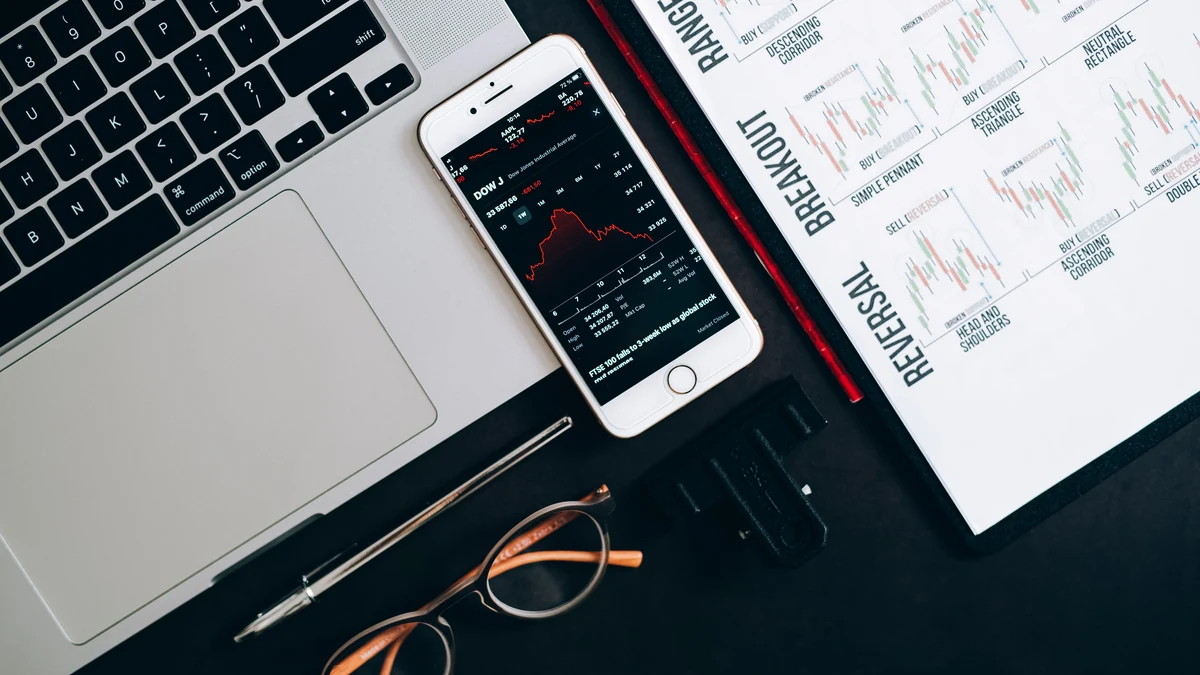 Laptop, smartphone displaying a stock market chart, and printed financial charts on a desk.
