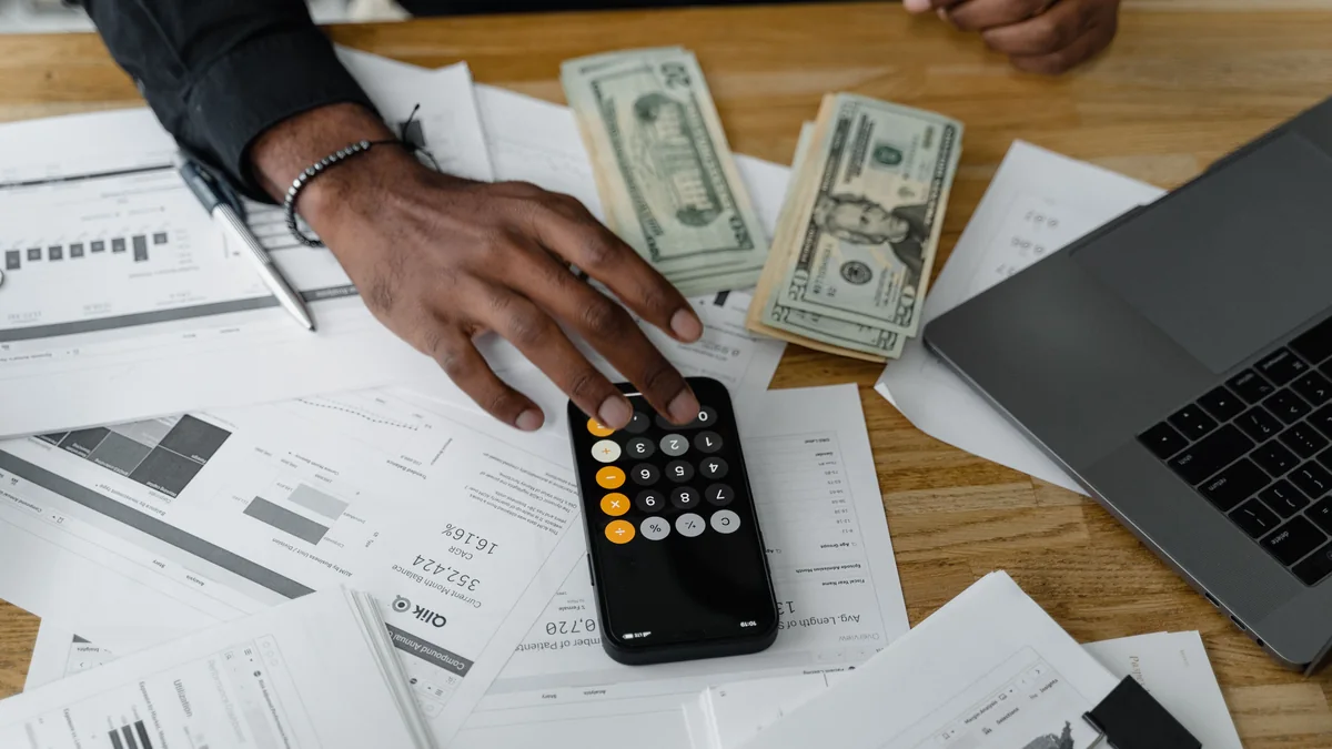 Hands using a calculator beside cash, financial documents and a laptop.