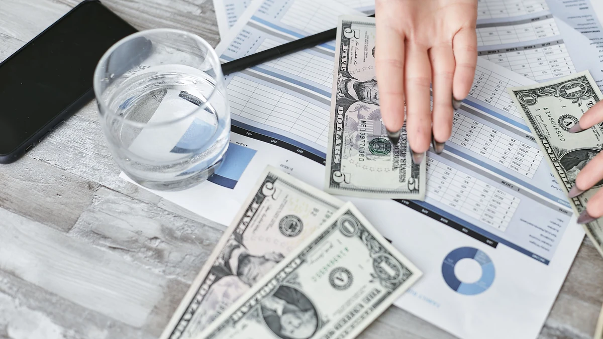 A hand above U.S. dollar bills and financial documents on a desk.