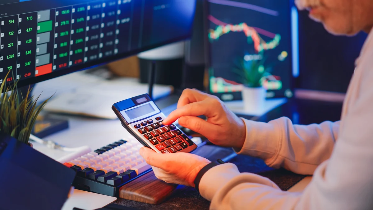 A person using a calculator at a desk with financial market screens in the background.