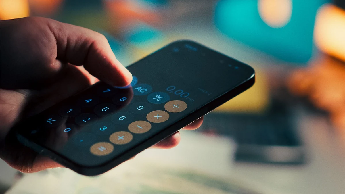 A hand using a smartphone calculator above cash and a notebook.