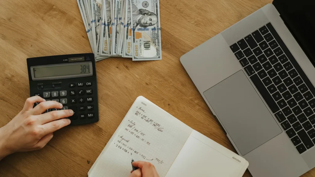 Hands using a calculator and notebook beside cash and a laptop for financial planning.