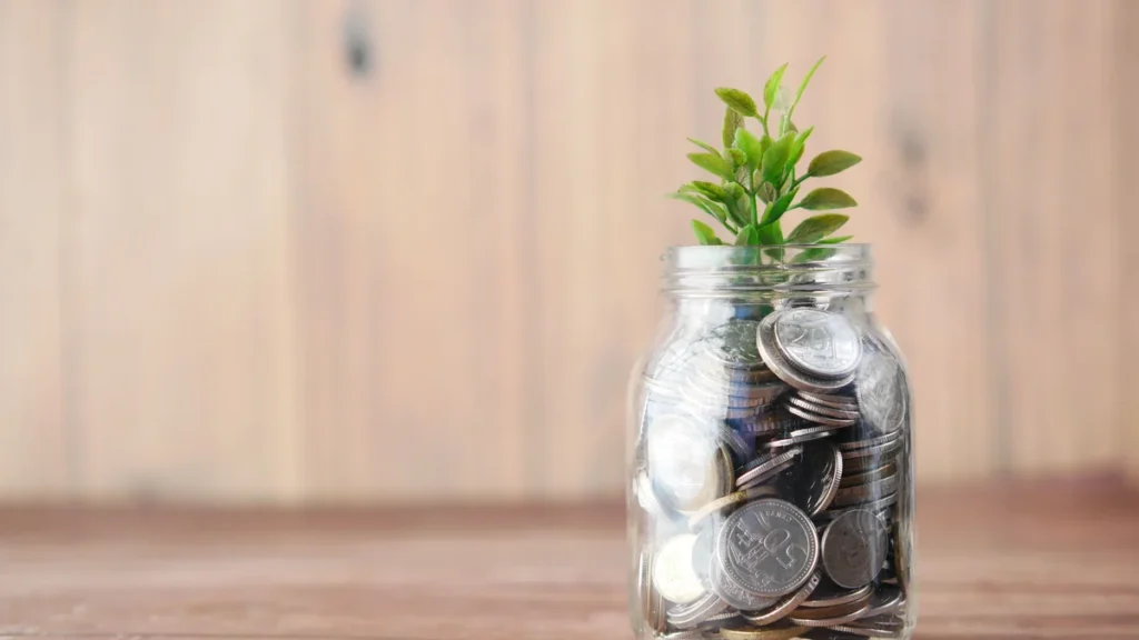 A glass jar filled with coins beside a small green plant.