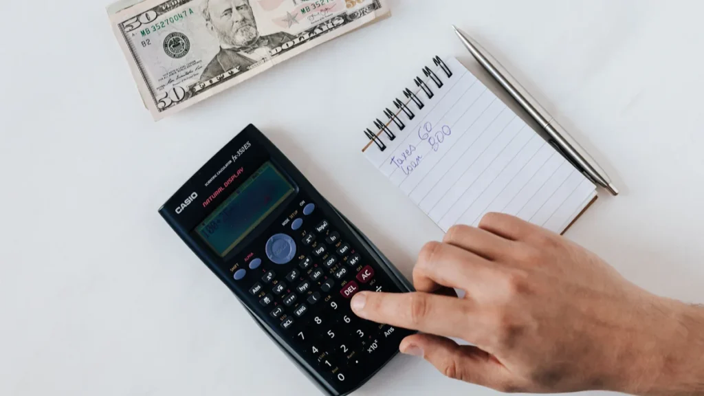 A hand using a calculator beside cash and a notebook during personal financial planning.