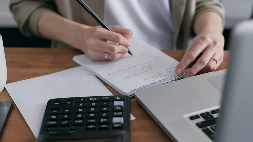 A person writing financial calculations in a notebook beside a calculator and laptop.