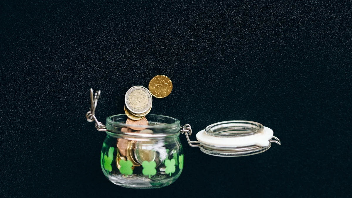 Coins falling into an open glass jar against a dark background.