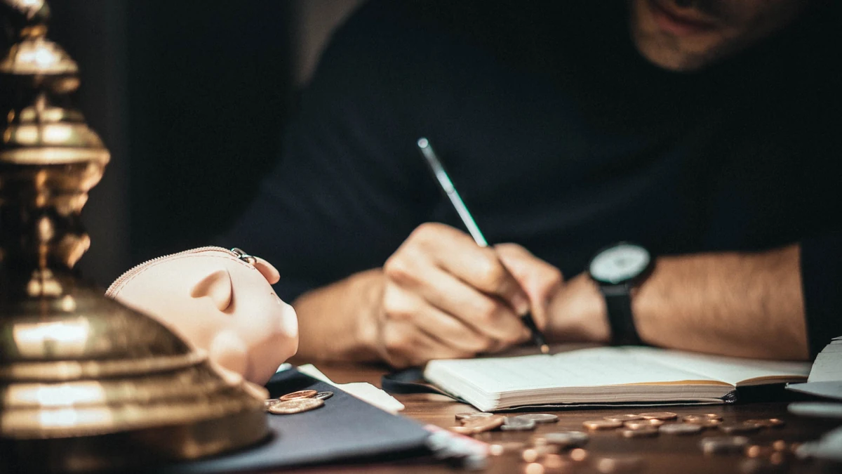 A person writing in a notebook beside a piggy bank and scattered coins on a desk.