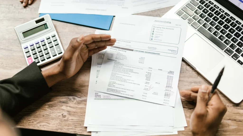Hands reviewing financial documents with a calculator and laptop on a desk.