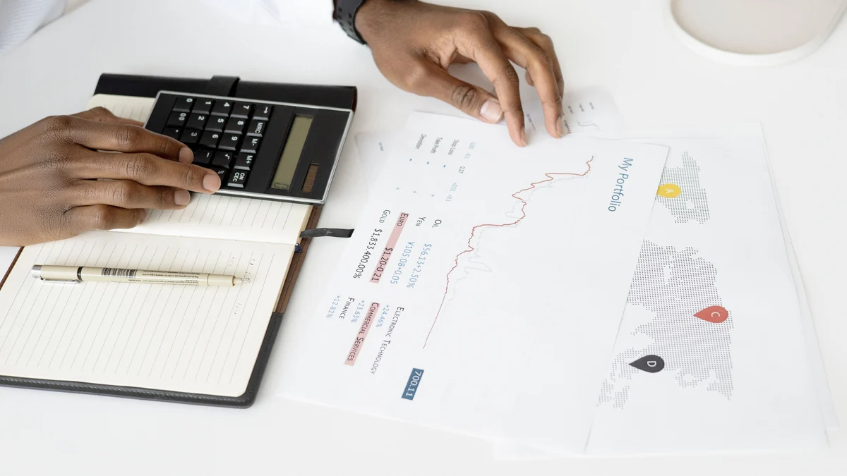 Hands using a calculator beside printed portfolio charts and a notebook on a desk.