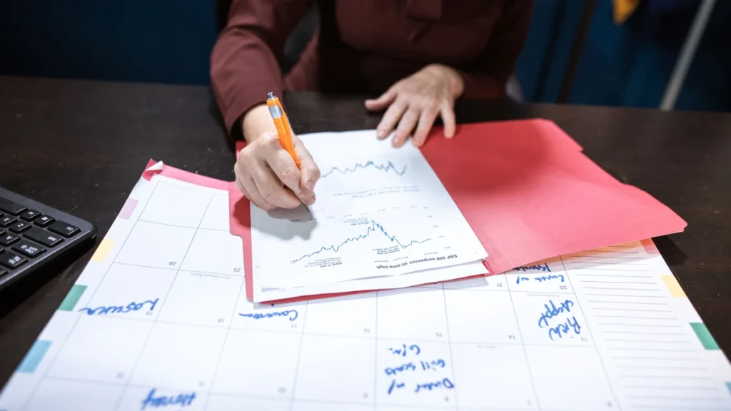 A person reviewing financial charts beside a monthly calendar on a desk.