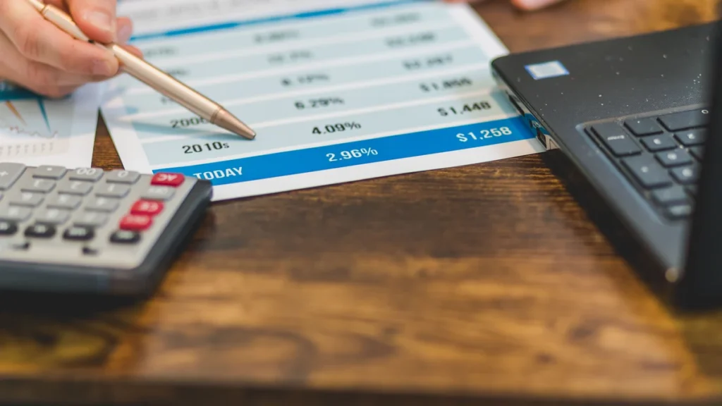 Hands using a pen to review interest-rate figures beside a calculator and laptop.