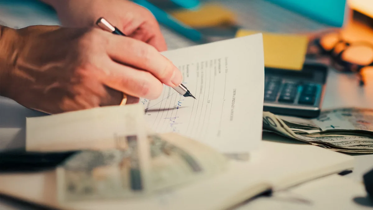 Person signing financial paperwork beside a calculator and cash on a desk.